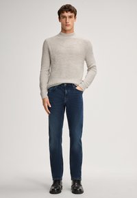 Maglione lavorato a maglia grigio chiaro con un collo alto, abbinato a jeans in denim blu scuro. Il modello indossa stivaletti neri in pelle.