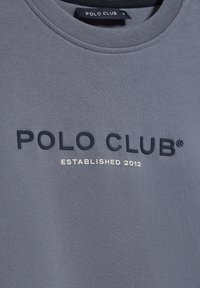 Grå sweatshirt med rund halsringning. Har en texturerad svart logotyp "POLO CLUB" och "ETABLERT 2012" i guld nedanför. Mjuk tyg.