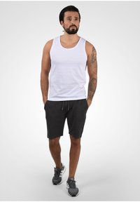 Débardeur en coton blanc associé à un short de sport gris foncé. La mannequin porte des baskets grises avec des accents blancs. Design simple et ajusté.