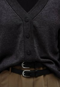 Cardigan gri închis cu decolteu în V, având cinci nasturi negri și margini cu tiv, asortat cu o curea neagră deasupra pantalonilor plisati călduți de culoare bej.