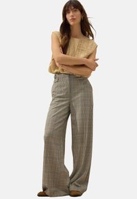 Beige ärmelloses Oberteil mit strukturierten Details; weit geschnittene Karohosen in Braun, Creme und Grün; neutralfarbene Schuhe.