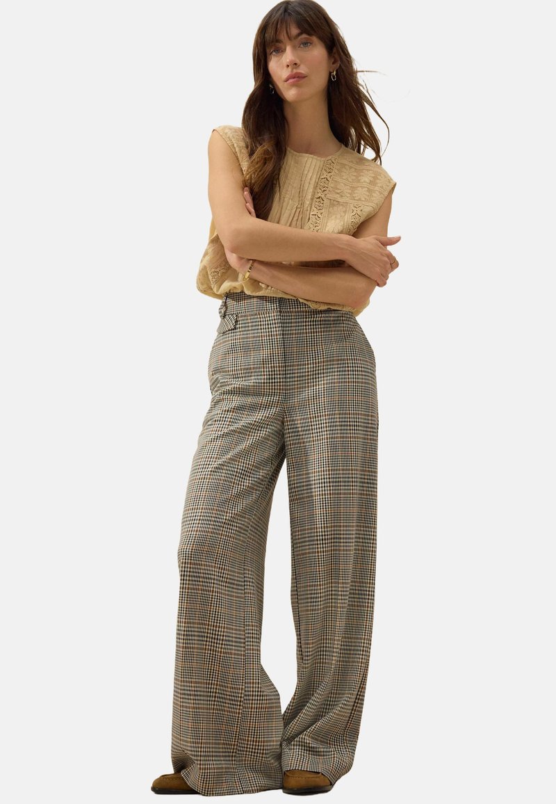 Beige ärmelloses Oberteil mit strukturierten Details; weit geschnittene Karohosen in Braun, Creme und Grün; neutralfarbene Schuhe.