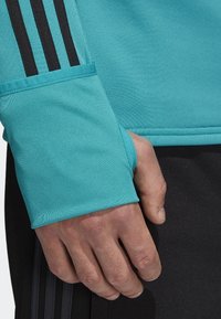 Turkusowa bluzka sportowa z czarnymi paskami na rękawach. Materiał wydaje się teksturowany, z widocznymi szwami i otworem na kciuk.