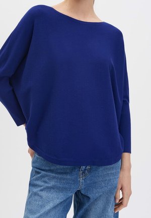 Persoon draagt een loszittende, donkerblauwe top met lange mouwen en een brede halslijn en lichtblauwe spijkerbroek.