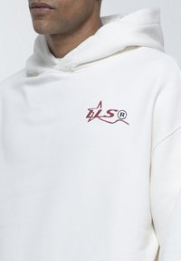 Uomo che indossa un hoodie bianco con una piccola stella bordeaux e le iniziali "LS" nel petto.
