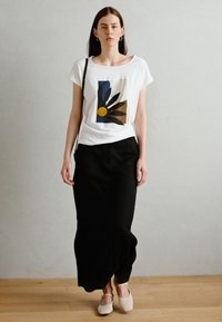 T-shirt blanc à manches courtes avec un design floral abstrait coloré ; associé à une jupe noire longue et des chaussures de couleur claire.