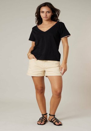 Femme debout portant un haut noir à manches courtes avec col en V, un short beige et des sandales plates à lanières noires sur un fond uni.