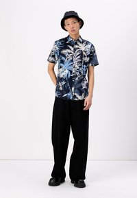 Chemise à manches courtes bleu marine avec un imprimé de feuillage tropical blanc et bleu clair, associée à un pantalon large noir et des chaussures noires épaisses.