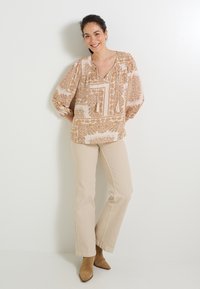 Blusa de color rosa claro con un patrón de paisley en marrón, con escote en V y mangas largas abullonadas. Combinada con pantalones acampanados beige y botas de tobillo marrones.