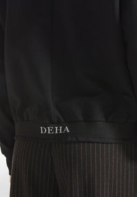 Dettaglio della parte inferiore di una felpa nera con la scritta bianca "DEHA" su pantaloni marroni a righe.