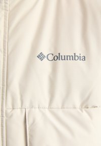 Bēžs Columbia apģērbs ar gludu tekstūru, ar tumši pelēku logo. Dizainā iekļautas horizontālas šuves un minimālisma stils.