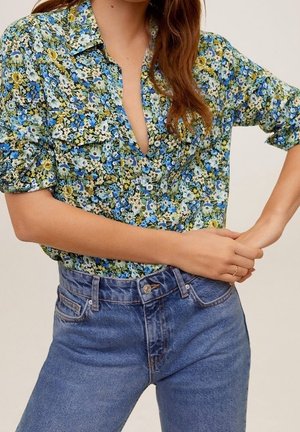 Frau trägt ein blau-grün-gelbes, geblümtes Hemd mit aufgekrempelten Ärmeln und blaue hochtaillierte Jeans, die Hände an der Taille verschränkt.