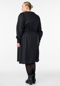 Frau mit blonden Haaren im Dutt trägt ein schwarzes, langärmliges, tailliertes Kleid, schwarze Strumpfhosen und schwarze Stiefeletten und steht nach links gewandt.