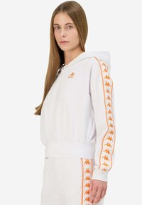 Sudadera blanca con cremallera y logo naranja de Kappa, además de franjas en las mangas. Fabricada con un material suave, cuenta con capucha y bolsillos laterales.