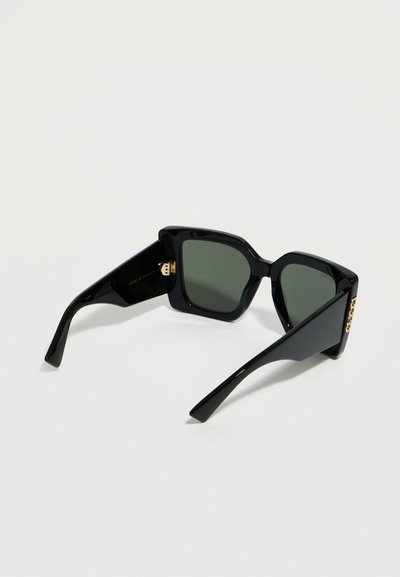 Μαύρα υπερμεγέθη γυαλιά ηλίου με τετράγωνο πλαίσιο και σκουρόχρωμους φακούς, με χρυσή λεπτομέρεια λογότυπου στις βραχίονες, που ακουμπούν σε φωτεινή επιφάνεια.