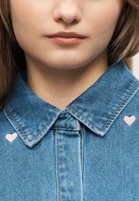 Primer plano del rostro inferior y cuello de una mujer que lleva una camisa de mezclilla azul con pequeños corazones bordados en rosa cerca del cuello.