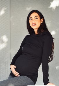 Top nero a maniche lunghe con collo a cowl e tessuto elasticizzato, indossato sopra un ventre visibilmente incinta. La modella ha i capelli lunghi e indossa leggings grigi.