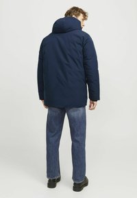 Doudoune bleu marine avec capuche, manches longues et coupe décontractée. Associée à un jean bleu clair et des chaussures noires. Design simple et fonctionnel.