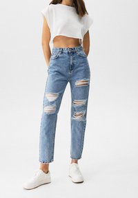 Jeans de tiro alto de color azul claro con desgarros desgastados, complementados con una camiseta corta blanca y zapatillas blancas. Ajuste informal y textura de denim.