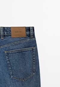 Dos d'un jean en denim bleu avec un patch de marque en cuir indiquant "Massimo Dutti" au-dessus de la poche droite sur un fond blanc.