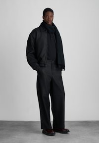 Schwarze Leder-Bomberjacke über einem schwarzen Polohemd, kombiniert mit weiten schwarzen Hosen. Ein schwarzer Schal umschmeichelt den Hals. Dunkelbraune Stiefel runden den Look ab.