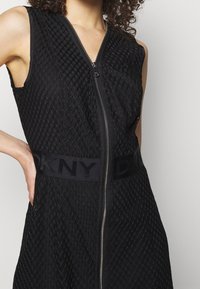 Robe noire à fermeture éclair sur le devant, en tissu texturé avec un motif en losange. Dotée d'une bande avec logo DKNY à la taille et d'un design sans manches.