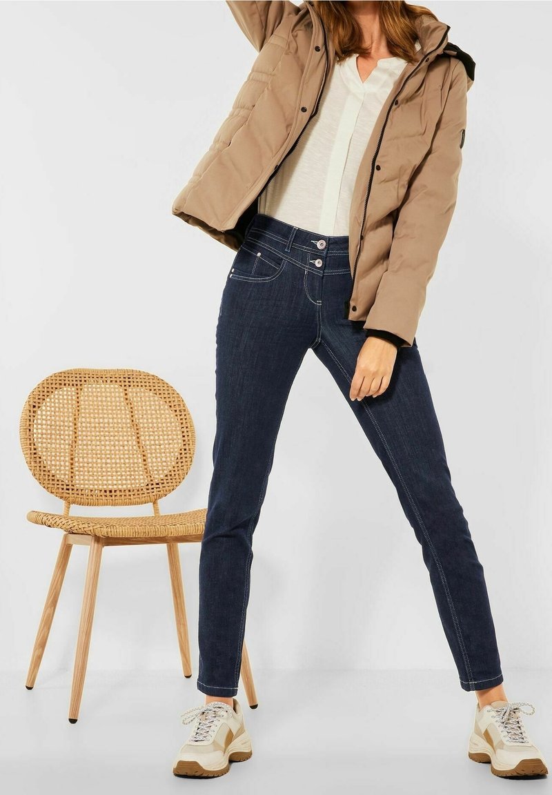 Femme portant une doudoune beige, un haut blanc, un jean bleu foncé et des baskets beiges, debout, jambes écartées à côté d'une chaise en bois tressé.