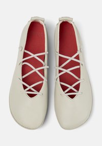 Valkoiset ballerinat, joissa on ristikkäiset nauhat ja punainen pohja, esillä harmaata taustaa vasten.