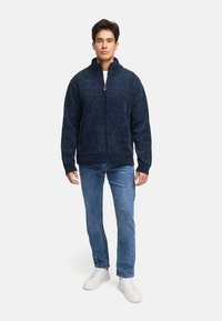 Marineblå strikket zip-sweater med høj krave, struktureret stof og lommer foran. Båret over en hvid skjorte og lyseblå jeans med hvide sneakers.