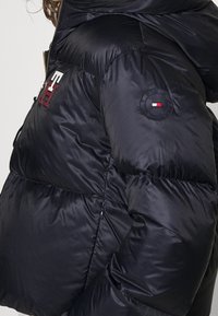 Sort pufjakke med en glansfuld finish, fremtrædende syninger, lynlås fortil og logo accenter på skulderen og brystet.