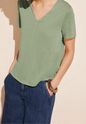 Person iført en lysegrøn V-hals kortærmet top og blå jeans med hænderne i lommerne og en brun armring på højre håndled.