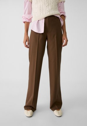 Persoon draagt bruine wijde broekspijpen, een roze overhemd met opgerolde mouwen, een crème gebreid vest en crème platte schoenen, staand tegen een effen achtergrond.