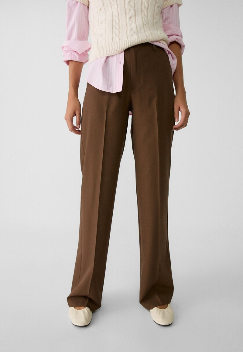 Personne portant un pantalon large marron, une chemise rose avec les manches retroussées, un gilet en maille crème et des chaussures plates crème, debout devant un fond uni.