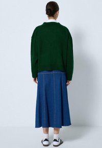 Groene gebreide trui met een ontspannen pasvorm, gecombineerd met een blauwe spijkerrok met een uitlopende snit en zichtbare stikseldetails.