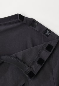 Σκουρό γκρι T-shirt με μαύρες ταινίες βέλκρο στις ραφές των ώμων, μαλακή υφή υφάσματος και ορατές ραφές γύρω από τον γιακά και τις άκρες.