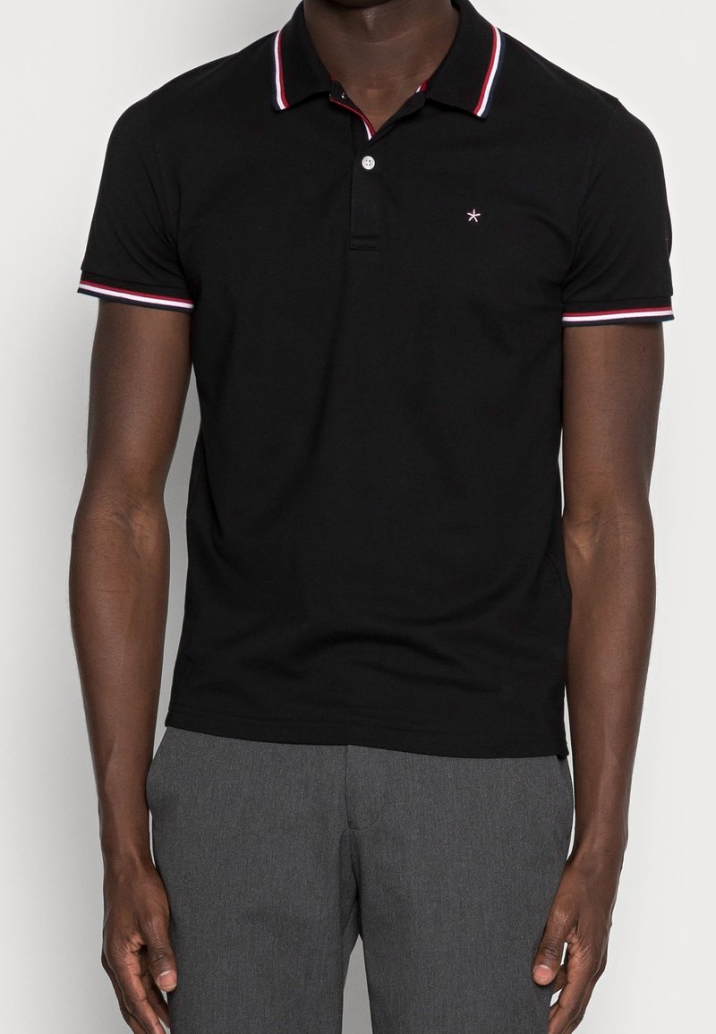 Polo noir à manches courtes avec col et poignets rayés rouge, blanc et noir, porté avec un pantalon gris.