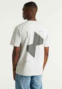 Korte mouwen, lichtgrijze t-shirt met een grote zwarte geometrische vorm en tekst op de achterkant, gemaakt van een zachte katoenmix.