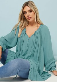 Tealblus med långa ärmar, plisserad design och knappstängning fram. Texturerad tyg med spetsdetaljer längs kanterna.