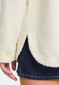 Weißer, flauschiger Mantel aus strukturiertem Stoff, mit einer Seitentasche, gelbem Knopfakzent und darunter sichtbaren dunklen Denim-Shorts.