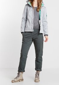 Vaaleanharmaa pehmeäkuorijakku turkooseilla yksityiskohdilla, harmaa T-paita, tummanvihreät housut ja vaaleanruskeat vaelluskengät. Sisältää etutaskut ja vetoketjukiinnityksen.