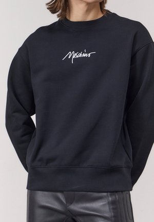 Schwarzer Pullover mit weißem "Moschino"-Logo, getragen mit schwarzen Lederhosen, zeigt Oberkörper und teilweise bedecktes Gesicht.