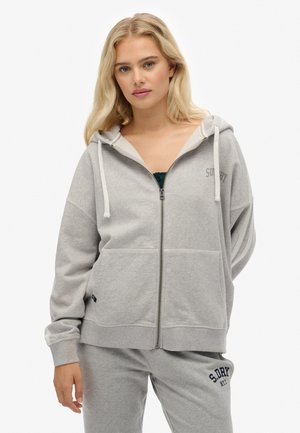 Blond kvinde iført en lysegrå zip-up hoodie og matchende sweatpants med "S.DRY" logo, stående mod en hvid baggrund.