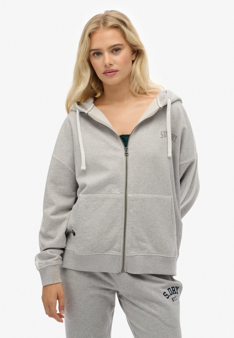 Mujer rubia vestida con una sudadera con cremallera gris claro y pantalones de chándal a juego con el logo "S.DRY", de pie frente a un fondo blanco.