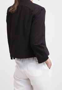 Femme portant un blazer noir court à manches trois-quarts et un pantalon blanc taille haute, mains dans les poches.
