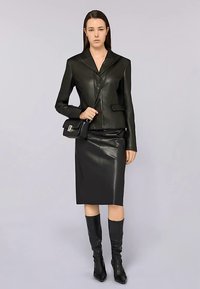 Zwarte leren blazer met piek revers en zijzak, gecombineerd met een aansluitende zwarte leren kokerrok en kniehoge zwarte laarzen. Minimalistische handtas.