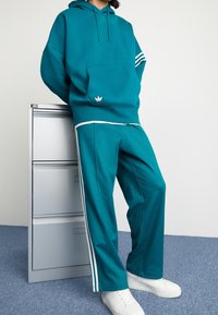 Turkos oversized hoodie med ficka fram och vita ränder på ärmarna, ihopparad med matchande turkosa byxor med sidostripes och vita skor.