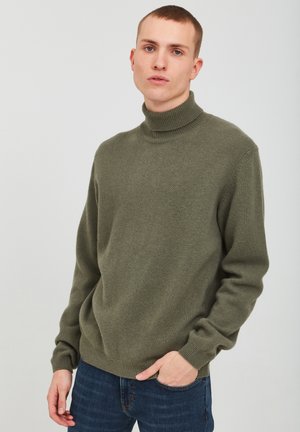 Suéter de cuello alto verde oliva hecho de una tela suave, con un corte relajado y puños acanalados. Se combina con unos jeans azules en un lavado oscuro.