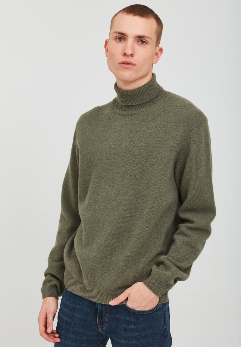 Maglione a collo alto di colore verde oliva realizzato in tessuto morbido, con una vestibilità rilassata e polsini a coste. Abbinato a jeans blu in un lavaggio scuro.