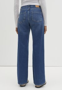 Jean en denim bleu à jambes larges, avec une taille haute, des poches arrière et un patch en cuir marron sur la ceinture arrière. Conçu pour un ajustement ample.