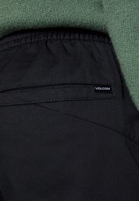 Gros plan sur la poche d’un pantalon noir avec taille élastique, une petite étiquette « Volcom » et un pull en maille vert au-dessus.